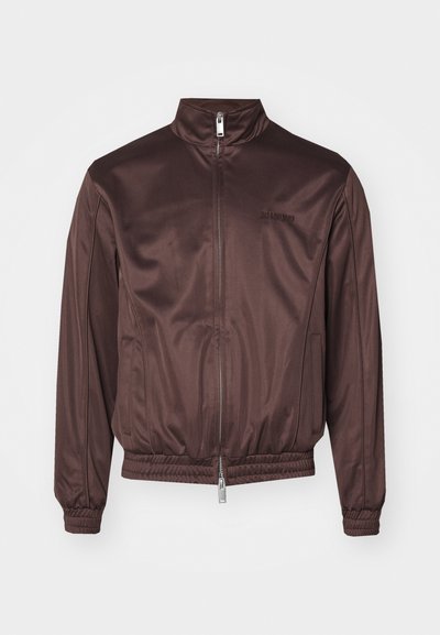 Jaqueta bomber castanha feita de tecido brilhante com colarinho alto, fecho frontal, punhos elásticos e faixa na cintura. Apresenta uma marcação mínima no peito.