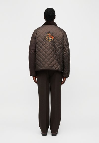 Άνδρας που φοράει σκούρο καφέ καπιτονέ μπουφάν με κεντημένο σχέδιο δράκου και το κείμενο «Barbour» στην πλάτη, σε συνδυασμό με καφέ παντελόνι.