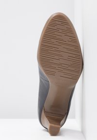 Chaussure à talon haut en cuir noir à finition lisse, semelle extérieure texturée de couleur beige avec un motif linéaire, et un accent en bois sur le talon.