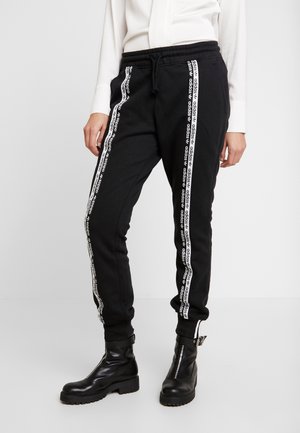 Personne portant un pantalon de survêtement Adidas noir avec des bandes blanches à logo sur les côtés et des bottines en cuir noir avec des fermetures éclair.