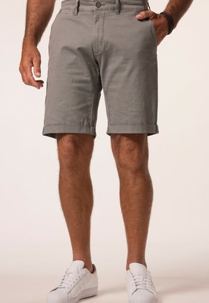 Mand iført grå shorts med opkrogede kanter, med den ene hånd i lommen og hvide sneakers, står foran en ensfarvet baggrund.