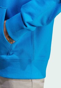 Person med en ljusblå sweatshirt och handen i fickan, som visar en liten tatuering på handleden.