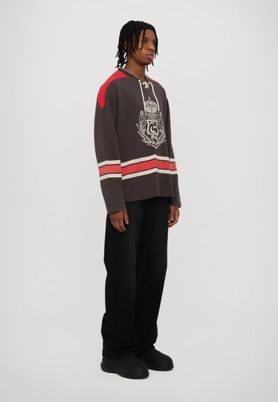 Sudadera gris de gran tamaño con acentos rojos, diseño gráfico, escote con cordones y rayas horizontales; combinada con pantalones negros y zapatos oscuros.