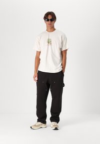 Camiseta de crema con diseño gráfico, chándal negro con bolsillos visibles y zapatillas beige con acentos grises y suelas texturadas. Gafas de sol oscuras.