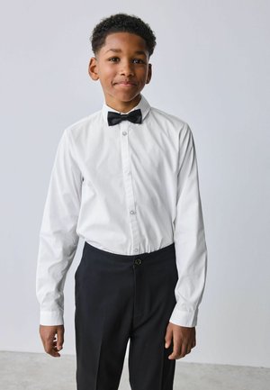Dreng iført hvid skjorte, sort butterfly og sorte bukser, stående mod en ensfarvet lys baggrund, smilende let.