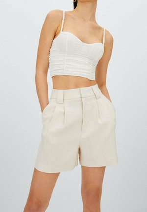 Haut court blanc en tissu texturé, présentant des bretelles fines et des fronces. Shorts taille haute beige clair avec des plis et des poches sur les côtés.
