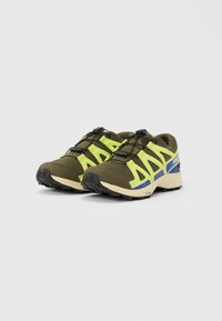 Ζευγάρι παπουτσιών για τρέξιμο σε μονοπάτια, ελιάς πράσινα και φωτεινά κίτρινα, με μαύρο σύστημα γρήγορου δέσιμου και μπεζ σόλα, γωνιακά στραμμένα ελαφρώς δεξιά σε λευκό φόντο.