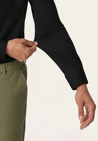 Schwarzes langärmliges Hemd mit einer taillierten, glatten Textur. Mit Bündchen versehene Ärmel und ein olivgrüner Bund, der im unteren Teil sichtbar ist.