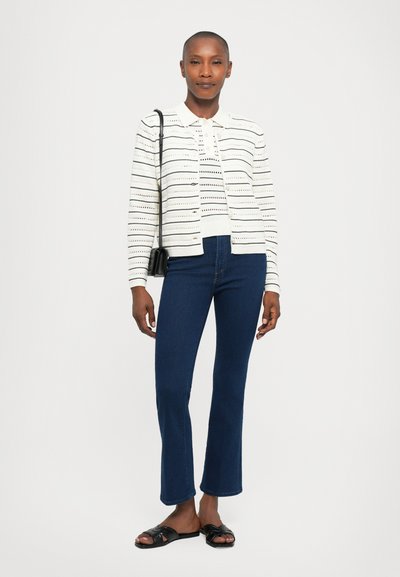 Witte cardigan met horizontale uitsnijdingen, gedragen met marineblauwe jeans en zwarte sandalen. Heeft een kraag en knopen. Compacte zwarte handtas.