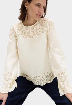 Kobieta w kremowej koronkowej bluzce z kwiatowymi wzorami na rękawach, dekolcie i brzegu, ubrana w ciemne spodnie, siedząca z rękami spoczywającymi na kolanach.