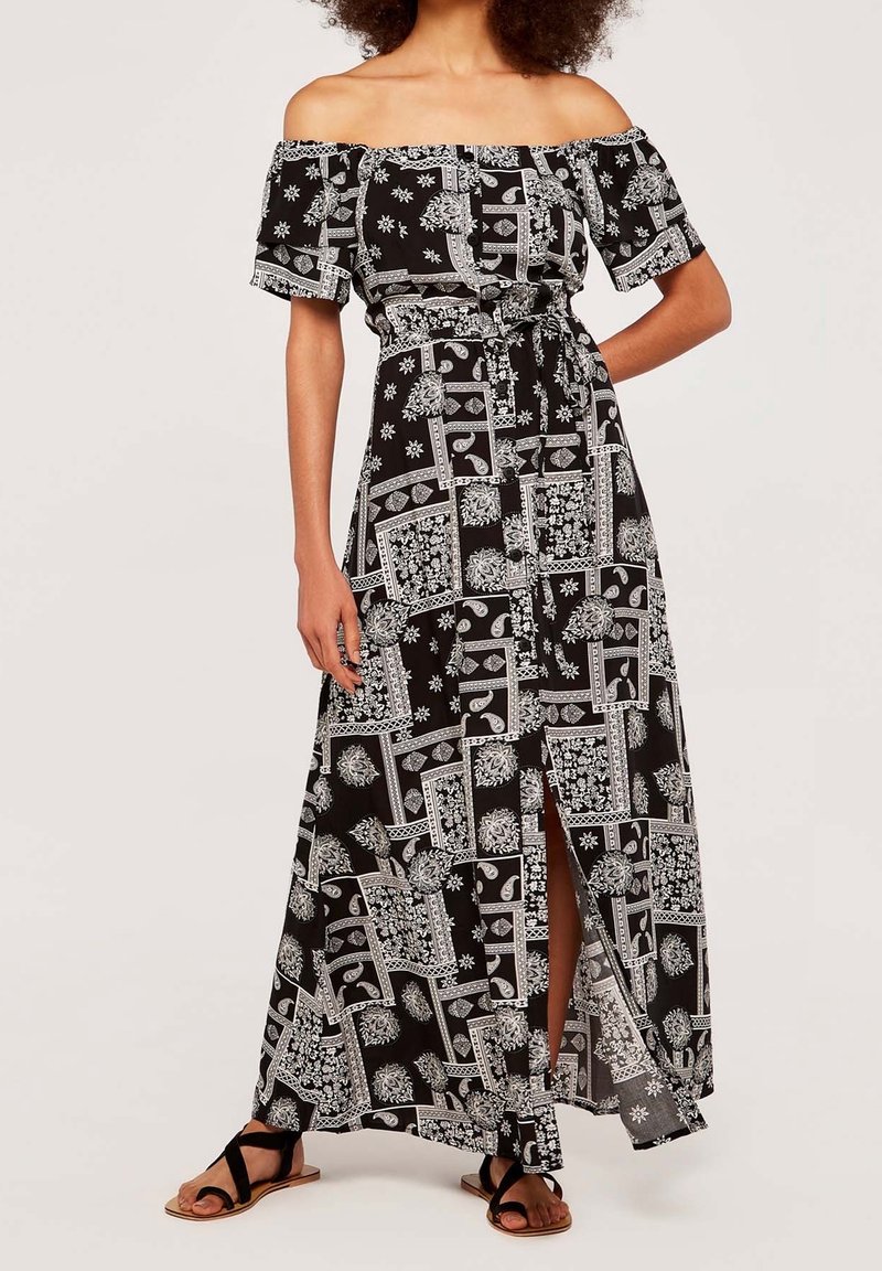Femme portant une robe maxi épaules dénudées noire et blanche avec motif floral et géométrique, fente à l'avant, taille nouée, et sandales noires à brides.