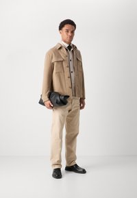 Veste beige avec de grands boutons et deux poches poitrine, associée à une chemise claire, un pantalon beige et des chaussures noires, tenant un sac en cuir noir.