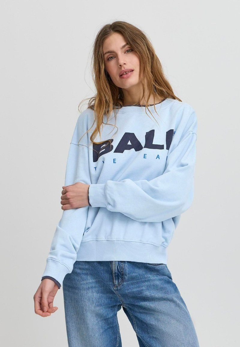 Lyseblå hettegenser med ribbestrikkede cuffs og midjebånd, med fet svart tekst "BALI" og mindre tekst under. Bært med blå jeans.