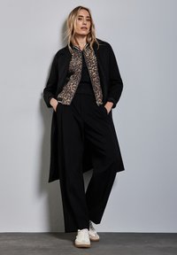 Manteau noir avec une veste bomber imprimé léopard sur un haut noir, associé à un pantalon noir à jambes larges et des baskets crème sur un fond gris.