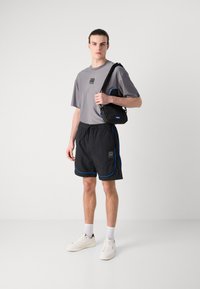Camisa gris de manga corta con logo, pantalones cortos negros con ribete azul, zapatillas blancas y una bolsa de hombro negra con acentos azules.