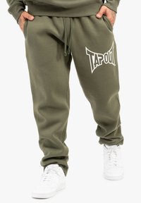 Olivengrønne joggerbukser med elastisk talje, snoretræk og stort hvidt "TAPOUT" logo på venstre side, parret med hvide sneakers.