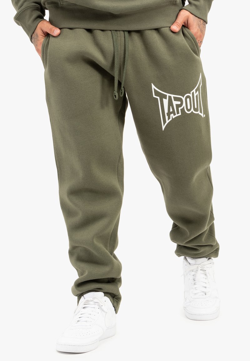 Olivengrønne joggerbukser med elastisk talje, snoretræk og stort hvidt "TAPOUT" logo på venstre side, parret med hvide sneakers.