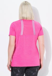 Persona con capelli biondi che indossa una maglietta sportiva a maniche corte di un vivace colore rosa con strisce riflettenti sul retro e leggins neri a fantasia.