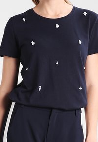 Námořnická modrá bavlněná tričko s kulatým výstřihem a rozsypanými bílými perličkami na přední straně. Jednoduchý, uvolněný střih.