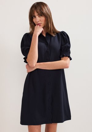 Vrouw met lichtbruin haar, gekleed in een zwarte jurk met knopen en opgeblazen korte mouwen, poseert met één hand die haar kin raakt en de andere arm gekruist.