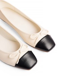 Ballerines en cuir beige avec un bout carré en vernis noir, ornées d'un petit nœud sur le devant. Texture lisse sur l'ensemble.
