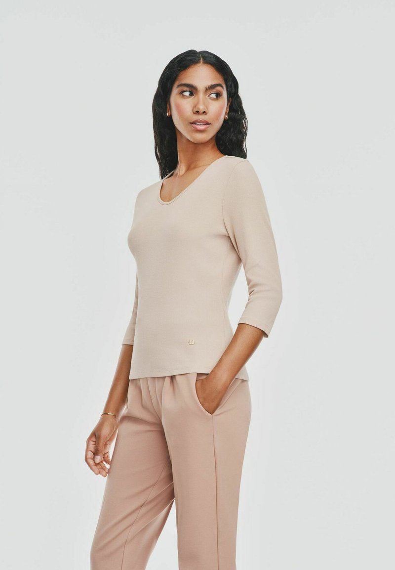 Jasno beżowa, prążkowana bluzka z długim rękawem, z okrągłym dekoltem i małym akcentem logo. Zestawiona z dopasowanymi luźnymi spodniami. Gładka faktura.