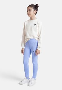 Νεαρή κοπέλα όρθια, φοράει λευκό φούτερ Nike, ανοιχτό μπλε κολάν Puma, λευκά αθλητικά παπούτσια, τα μαλλιά της σε διπλό κότσο, κοιτάζει προς τα δεξιά.