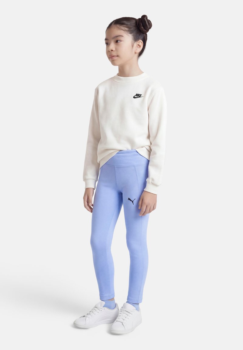 Νεαρή κοπέλα όρθια, φοράει λευκό φούτερ Nike, ανοιχτό μπλε κολάν Puma, λευκά αθλητικά παπούτσια, τα μαλλιά της σε διπλό κότσο, κοιτάζει προς τα δεξιά.