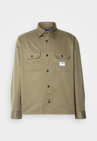 Chemise à manches longues vert olive avec deux poches à rabat sur le devant, boutons noirs et petit patch logo blanc sur la poche gauche.