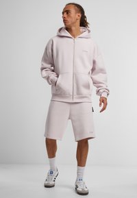 Lys rosa zip-up hettegenser med kengurulommer, kombinert med matchende shorts. Mykt stoff, avslappet passform og subtile merkevaredetaljer på begge plagg.
