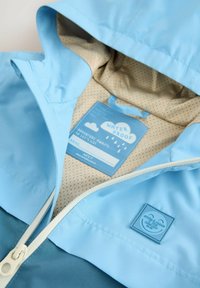 Giacca impermeabile in blu chiaro e verde acqua, con chiusura a zip e fodera beige perforata. Presenta un'etichetta con design di nuvole all'interno.