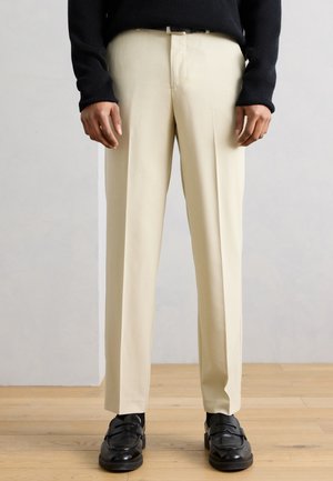 Άνδρας με μπεζ προσαρμοσμένο παντελόνι, μαύρη ζώνη, μαύρο πουλόβερ και μαύρα δερμάτινα μοκασίνια που στέκεται σε ξύλινο πάτωμα.