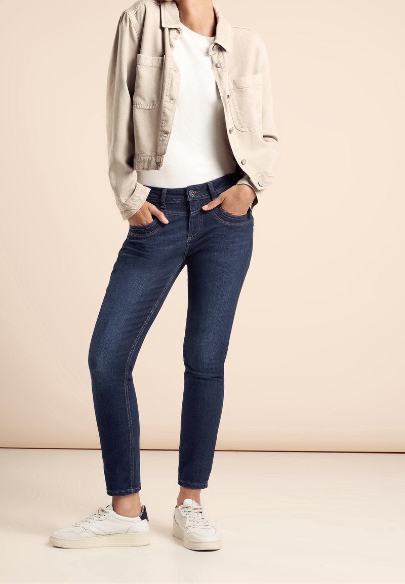 Personne portant un jean skinny bleu foncé, des baskets blanches, un haut blanc et une veste courte beige clair, les mains dans les poches.