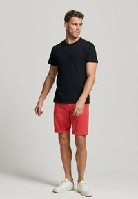 Camiseta negra de manga corta, pantalones cortos rojos y zapatillas blancas. La camiseta tiene un logo sutil, los pantalones cortos están doblados en el dobladillo para un ajuste relajado.