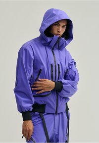 Lila kapucnis dzseki, könnyű anyagból készült, rugalmas mandzsettákkal, több cipzáras zsebbel és laza szabású kialakítással.