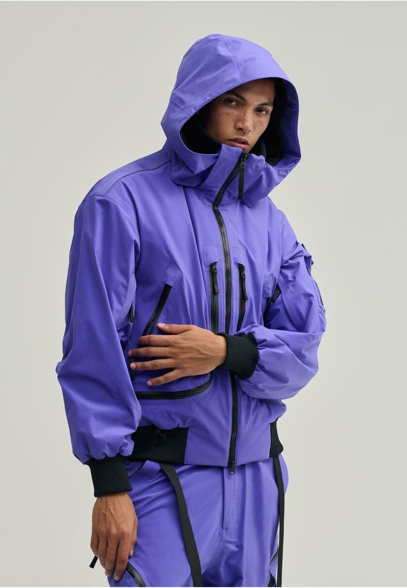 Lila kapucnis dzseki, könnyű anyagból készült, rugalmas mandzsettákkal, több cipzáras zsebbel és laza szabású kialakítással.