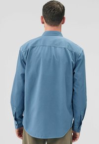 Een persoon in een lichtblauw, langslurf shirt staat met de rug naar de camera, waarbij de kraag en manchetten van het shirt zichtbaar zijn tegen een neutrale achtergrond.