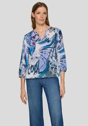 Frau trägt eine locker sitzende blaue Bluse mit abstrakten Mustern und dem Schriftzug „COOL“, kombiniert mit blauen Jeans, stehend vor einem schlichten Hintergrund.