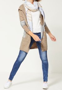 Béžový svetr s šedými rukávy, vrstvený přes bílý top. Modré džíny s úzkým střihem a bílé tenisky. Lehký pruhovaný šátek dodává detail.