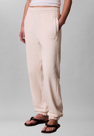 Persona que lleva puestos pantalones deportivos Calvin Klein de color beige claro con puños elásticos y bolsillos, combinados con sandalias negras tipo chancla.