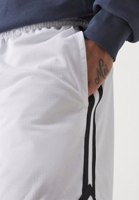  calções de malha brancos com acabamento texturizado, apresentando uma faixa lateral preta e uma cintura com cordão. Uma tatuagem é visível na mão.