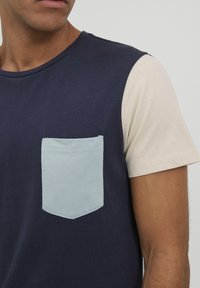 Tmavě modré tričko s krátkými béžovými rukávy, s světle modrou obdélníkovou kapsou na levé straně. Měkká bavlněná látka, jednoduchý design.