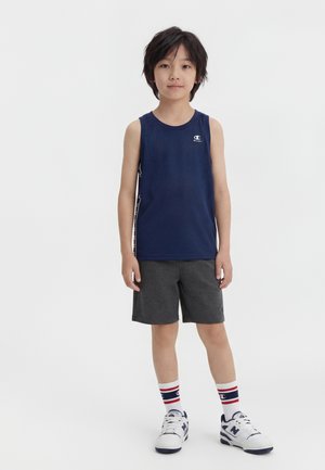 Fant, ki stoji, oblečen v temno modro brezzloženo majico, temno sive kratke hlače, bele športne nogavice z rdečimi in modrimi črtami ter bele superge.