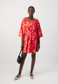 Punane lilleline kleit ruudukujulise kaelusega ja puhvainikega, rõhutatud vöökohalt. Kantud musta käekotiga ja heledate kingadega.