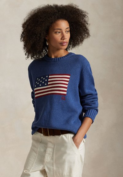 Frau mit natürlichen lockigen Haaren, die einen blauen Pullover mit eingestrickter amerikanischer Flagge und weiße Hosen trägt, schaut seitlich und hat eine Hand in der Tasche.