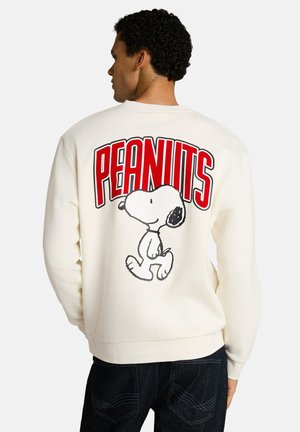 Mann mit lockigem Haar, der einen weißen Sweatshirt mit rotem Text "PEANUTS" und einer schwarz-weißen Snoopy-Grafik auf dem Rücken trägt.