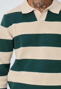 Polo en tricot présentant des rayures horizontales alternées vertes et beiges, avec un col classique et un détail de tissu texturé.