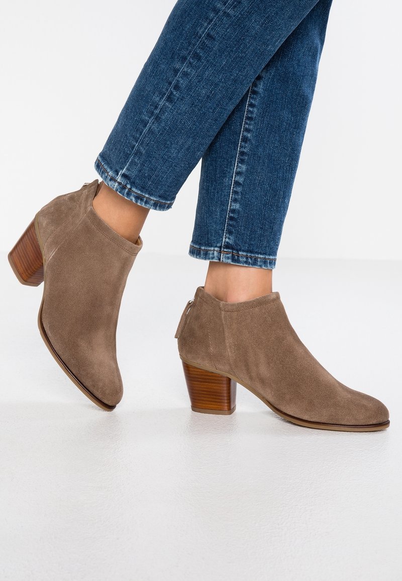 Bottes basses en daim marron avec un bout pointu, dotées d'un talon en bois superposé et d'une fermeture éclair à l'arrière. Portées avec un jean bleu.