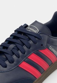 Jūrinės odinės sportinės bateliai su raudonomis juostelėmis. Yra tekstūruoti raišteliai ir apvalus logotipo ženkliukas. Smėlio spalvos guminis padas su briaunuotu raštu, užtikrinančiu sukibimą.