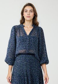Navy blauw, doorzichtige blouse met een V-hals, puffed sleeves en verspreide groene en witte stippen. Zachte textuur, lichtgewicht stof.